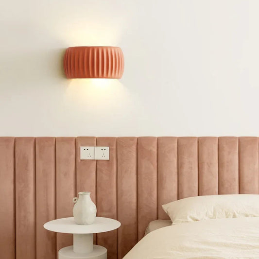 A modern bedroom scene featuring a ribbed terracotta orange wall sconce mounted on a white wall above a dusty rose velvet headboard. A small white side table with a white vase sits next to a bed with cream bedding.